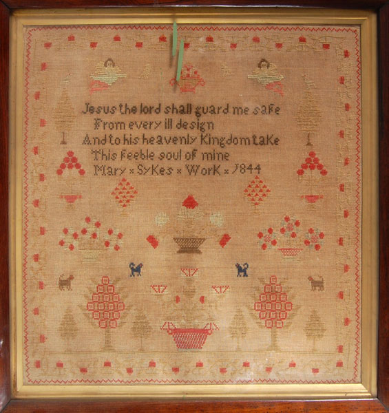 A framed sampler signed Mary Sykes 1844. Religious text in centre surrounded by flowers, trees, dogs, two cherubs and bordered by flowers.