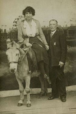Woman sat on a donkey with an older gentleman standing beside her. A studio photo with a backdrop featuring a Blackpool scene.