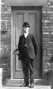 Man wearing a bowler hat standing in front of a door with drinks prices on a board on the left of the image.