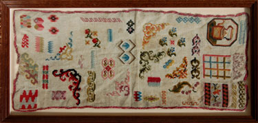 A framed sampler consisting of different designs dotted around; including a kettle, flowers, leaves, boxes. Edged with pink textile. Embroiderer unknown,