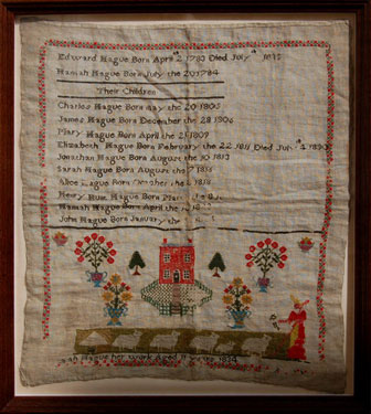 A framed sampler signed Sarah Hague, 1834. Memorial to Edward Hague and Hannah Hague and their ten children. Includes picture of a house, flowers, shepherdess and flock of sheep, with floral border.