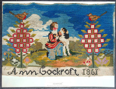 A framed woolwork sampler signed Ann Cockcroft, 1861. Child with dog, plus robins perched on top of two bushes.